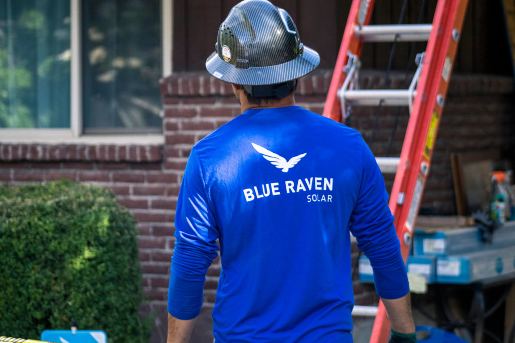 Blue Raven Solar installer in blue branded long-sleeve shirt and hardhat on, walking towards a ladder leading to the roof of a house