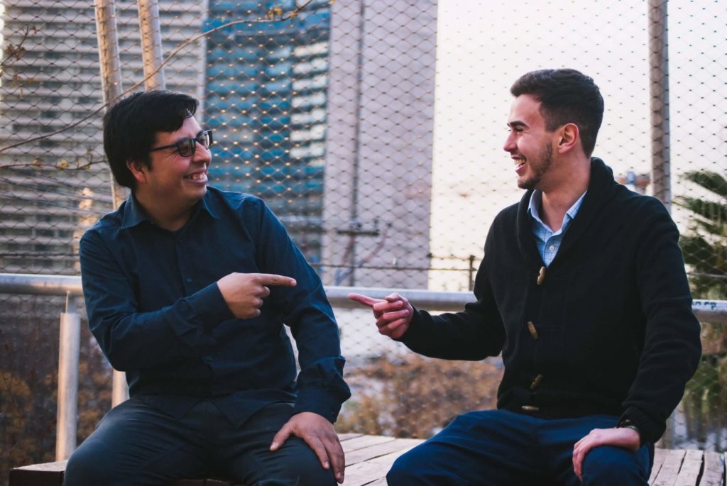 Two males in long-sleeve shirts and sweaters, smiling, and pointing at each other while sitting outside