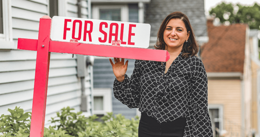 Female in black and white business casual attire standing behind a red house "For Sale" sign