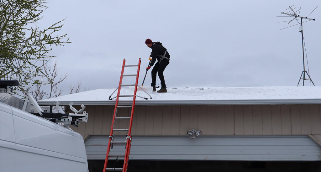 Male standing on roof with professional cleaning tools, clearing off snow and debris