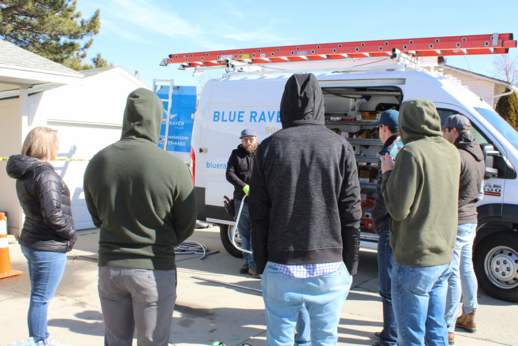 Small group of Blue Raven Solar employees on-site listening to instructions, in a semi-circle with a company branded install van in the background