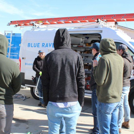 Small group of Blue Raven Solar employees on-site listening to instructions, in a semi-circle with a company branded install van in the background