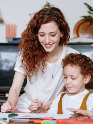 Red, curly haired female helping red-haired young child draw and color with markers