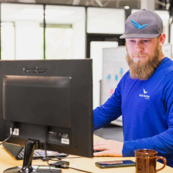 Male in Blue Raven Solar blue long sleeve shirt and branded hat, standing while looking at a computer monitor