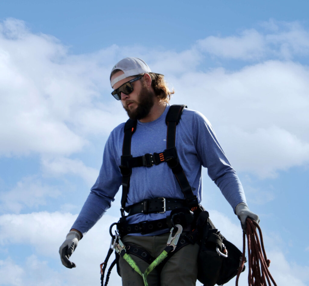 Blue Raven Solar installer in a harness, carrying specialized rope and equipment with blue skies and clouds behind