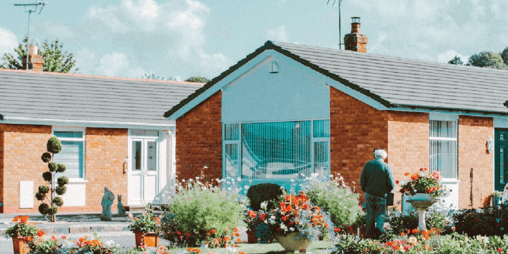 Male watering flowers outside of an orange, red brick, free-standing, single-level home