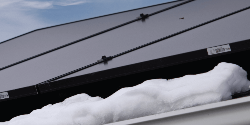 Solar panel array installed on roof with snow piles below and blue skies above