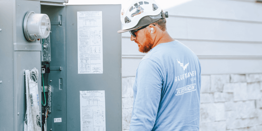 Blue Raven Solar installer in a branded light-blue shirt and a hard hat, evaluating electrical panel