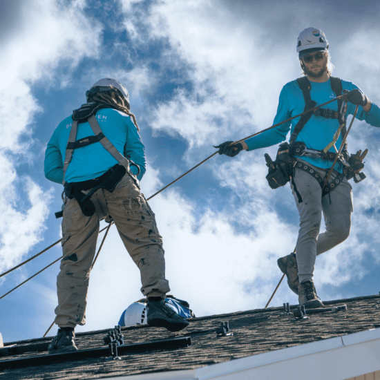 Two Blue Raven Solar installers in teal branded t-shirts utilizing specialized equipment to secure solar panels to a homeowner's roof