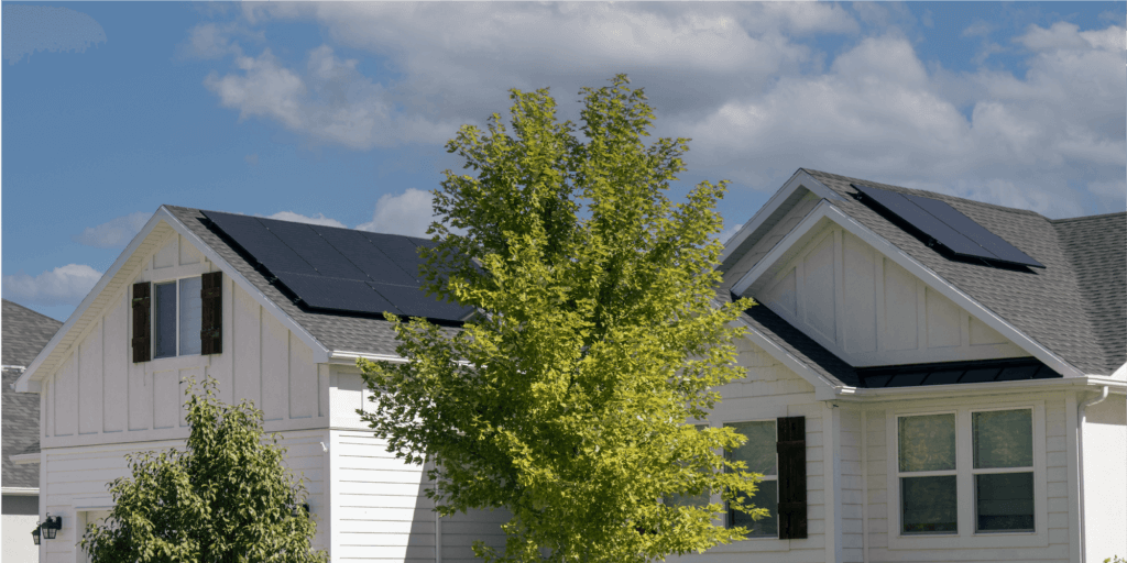 A large solar panel installation on a newly built white house with black trim, surrounded by lush green trees