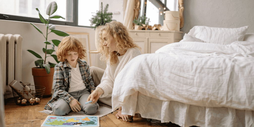 A mother is helping her son put together a puzzle of the continents of the world on the floor of a bedroom